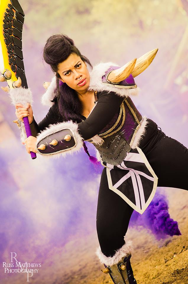 Woman wearing purple with horns and black clothing swings a sword made of lava rocks
