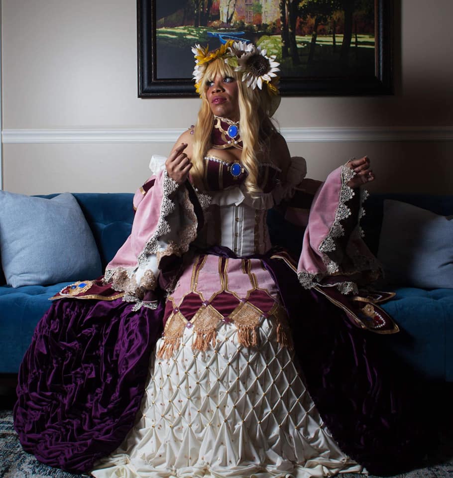 Juliette sits on a couch wearing a flower crown, neck corset, and an intricate pink and purple ballgown