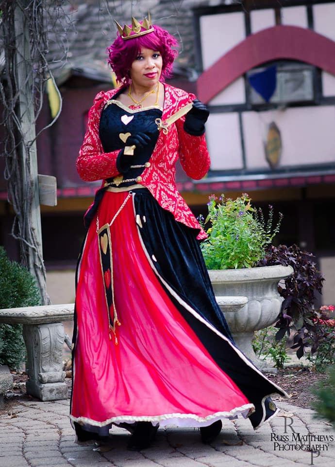 The Queen of Hearts grips a gold knife while wearing a crown, curly red hair, a gold and red jacket, black corset and a black and red gown with heart decorations.