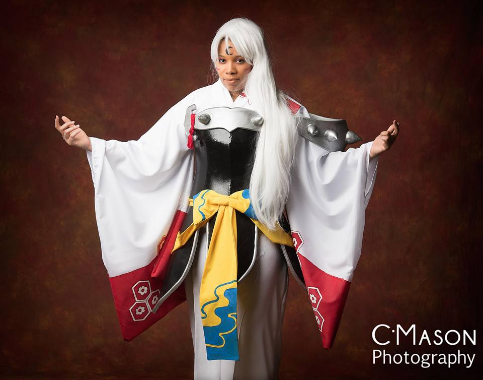 Sesshomaru stands in front of a brown background with his arms spread. Sesshomaru has long white hair, a red and white kimono and black and silver armor with a yellow and blue bow.