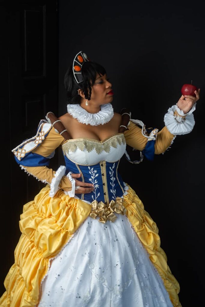 Snow White stands staring at a red apple in her hand while wearing an intricate blue and yellow ball gown.