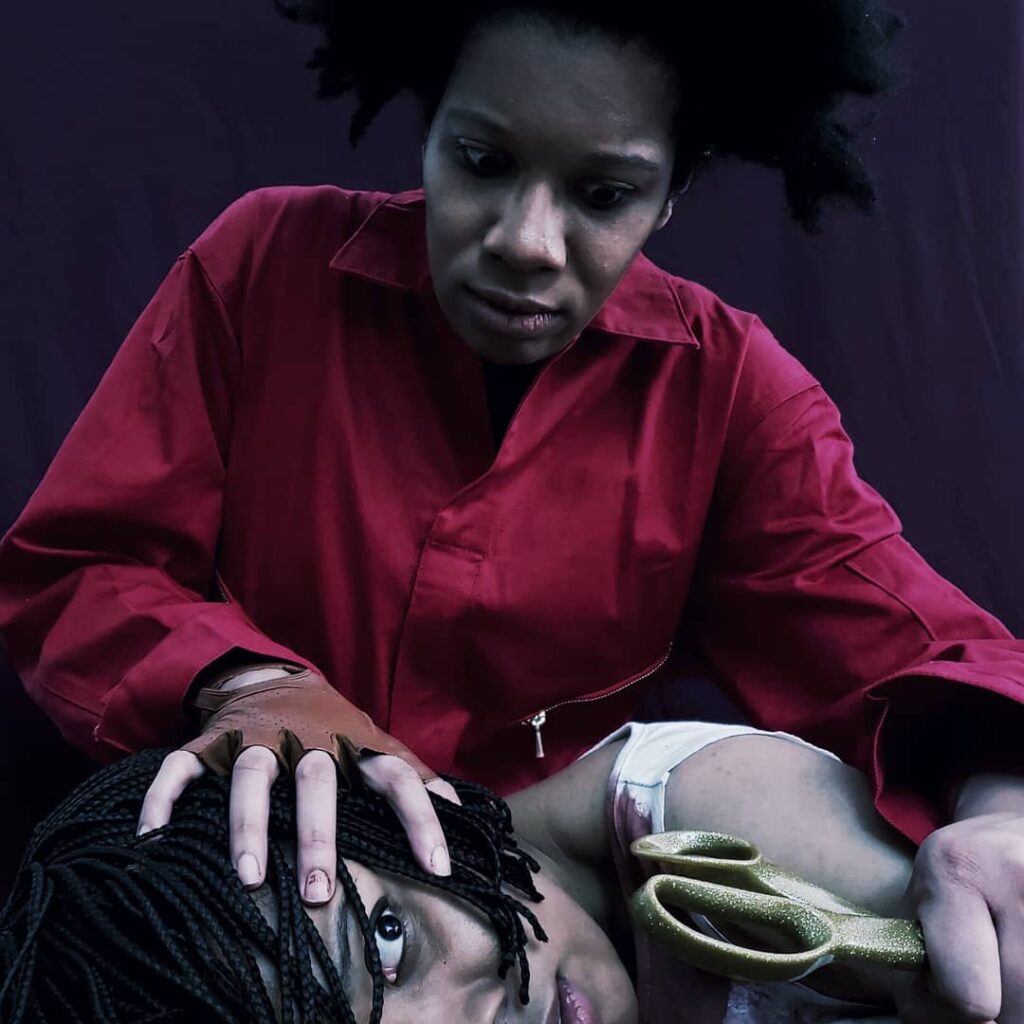 Red holds Adelaide's head down while holding a pair of gold scissors. Red wears natural hair, a red jump suit and a glove. Adelaide wears a bloody white shirt.