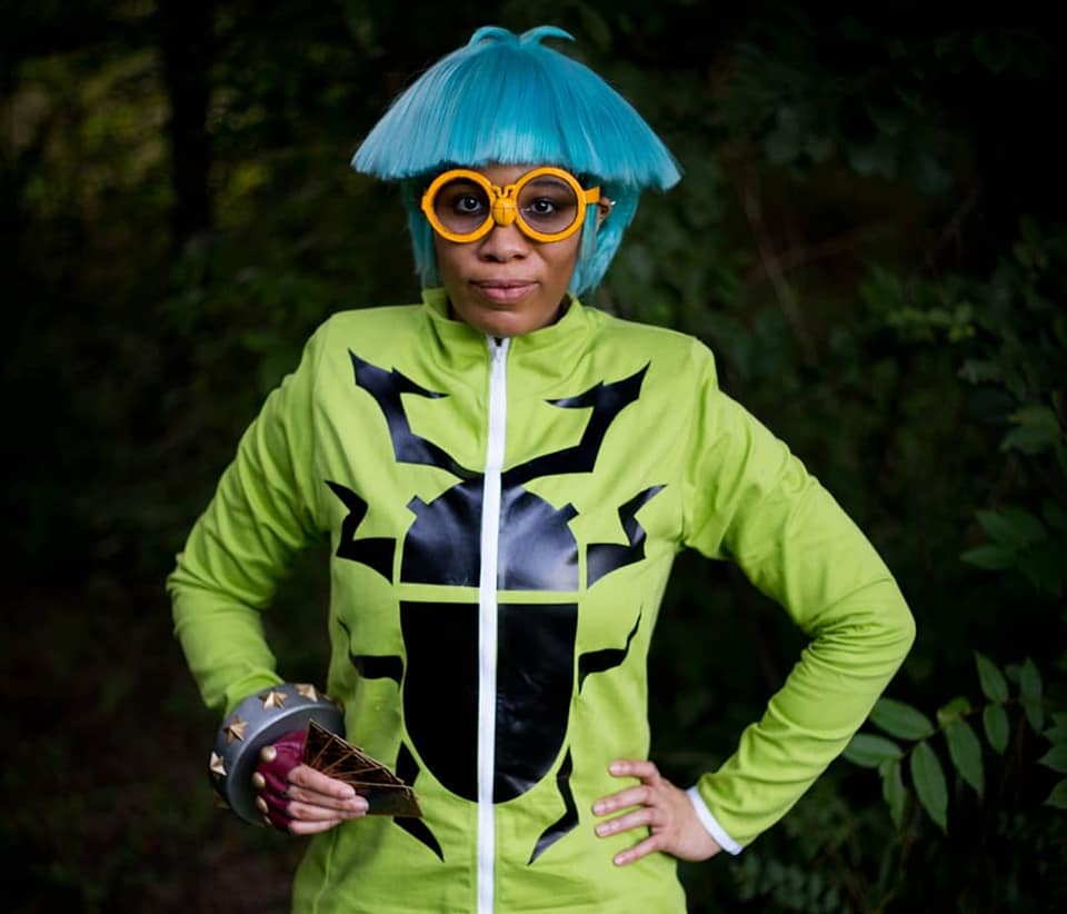 Weevil Underwood stands in a forest wearing blue hair, yellow glasses, and a bright green turtle neck with a beetle decal on the front holds Yugioh Cards.