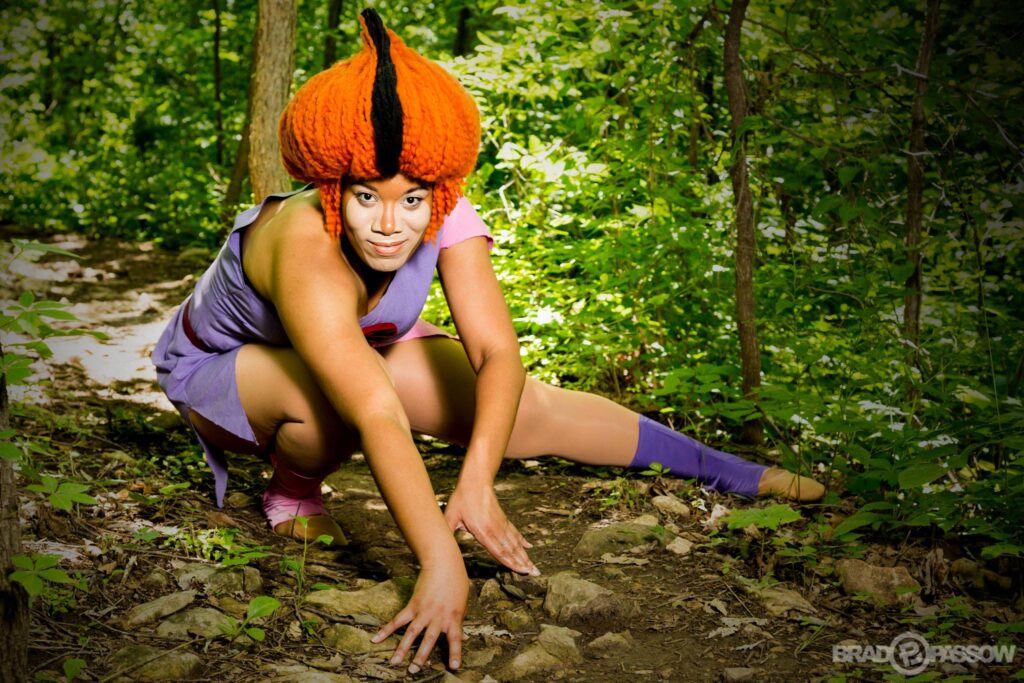 Wilykit is posed on the forrest floor. Wilykit has tall pointy orange and black hair and wears a pink and purple dress and pink and purple socks.